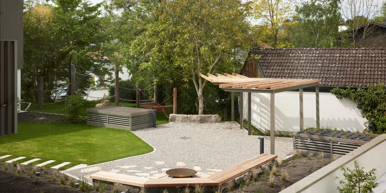 Außenbereich der VR Bank Andechs mit Sitzstufen, Feuerstelle, Pergola und grüner Landschaftsgestaltung.