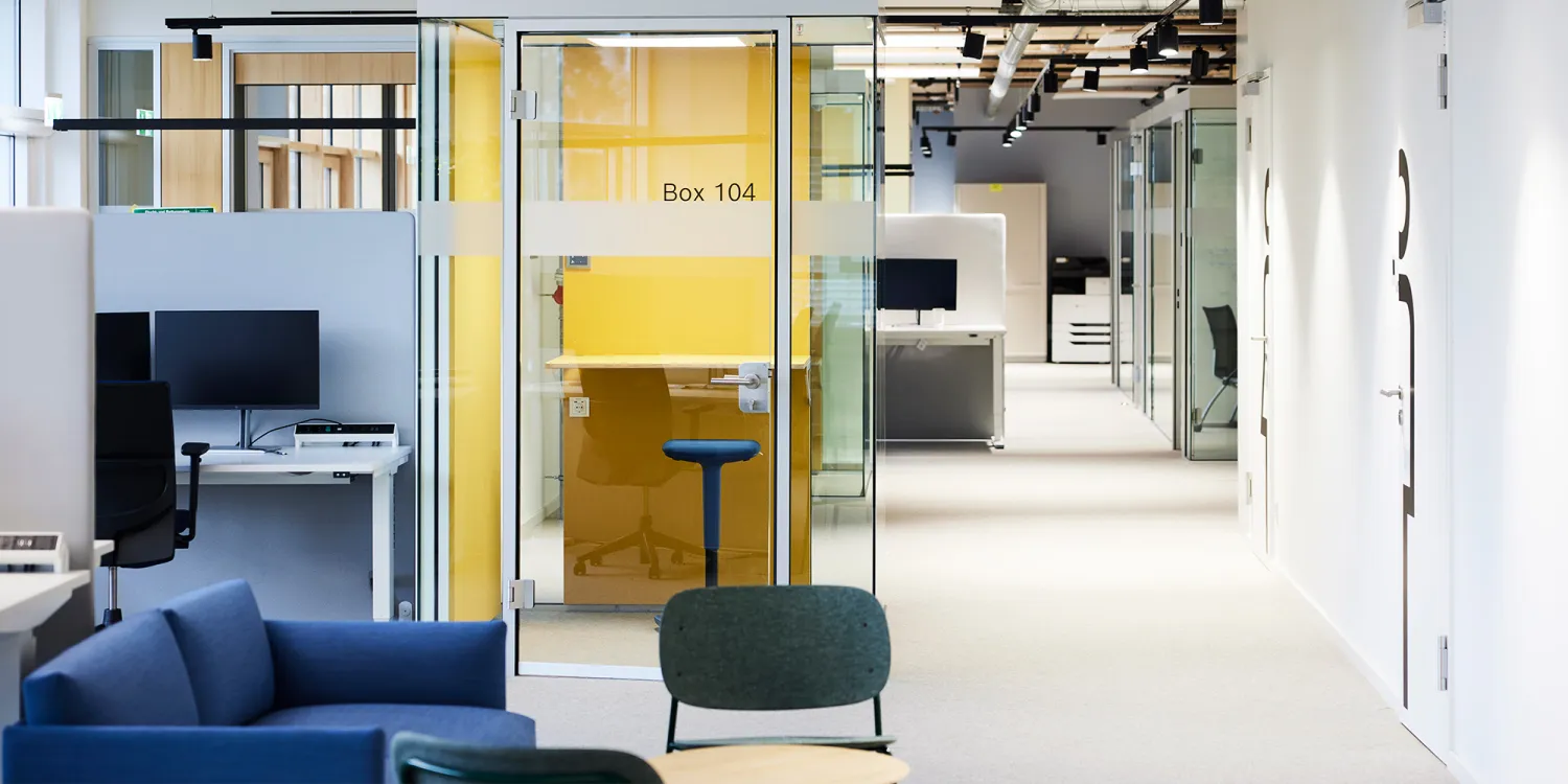 Bosch: Ein modernes Büro mit offenen Arbeitsplätzen, Glas-Meetingbox, gelber Akzentwand und Lounge-Sesseln.