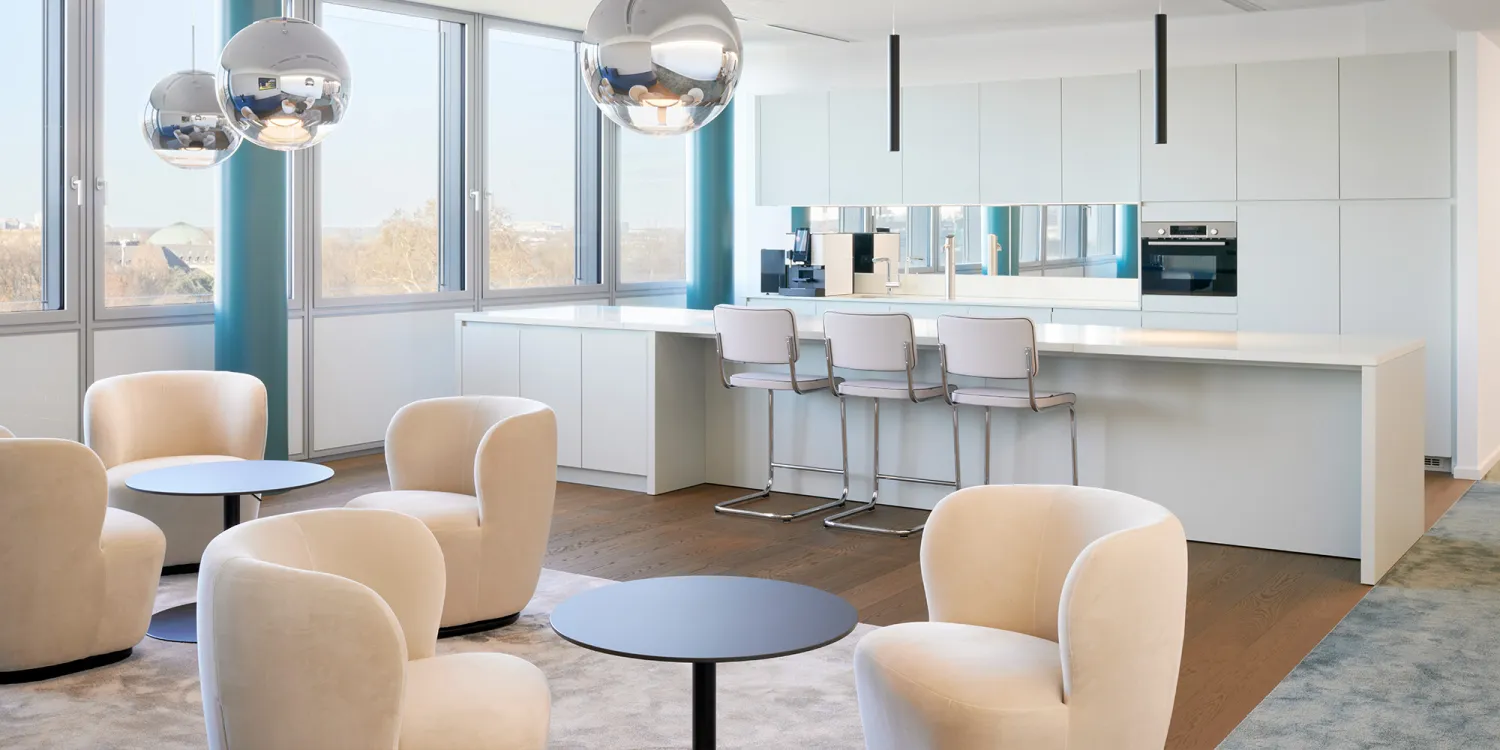 Hogan Lovells: Eine helle Lounge mit beigen Sesseln, schwarzen Tischen, einer modernen Küche und großen Fensterfronten.