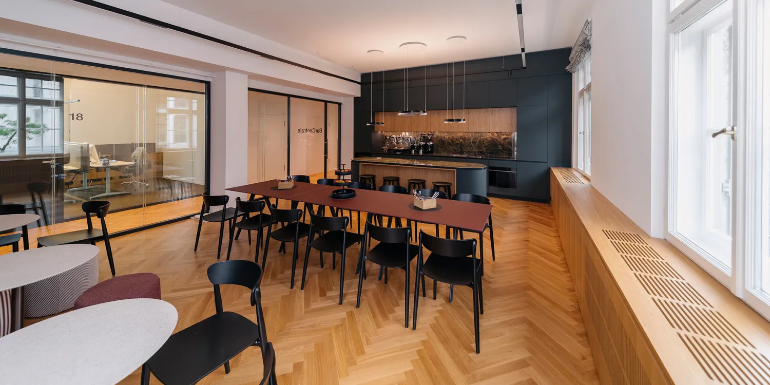 Offene Lounge mit großer Tafel, schwarzer Küche und Glasbüros im Hintergrund