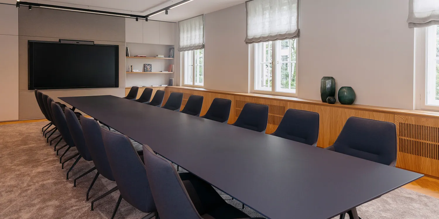 Großer Besprechungsraum mit einer langen Tafel und vielen Sitzmöglichkeiten. Im Hintergrund ist ein Screen