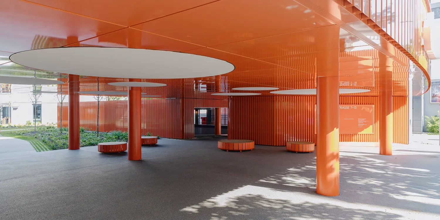 Elsenheimerstraße – überdachter Eingangsbereich mit leuchtend oranger Architektur und runden Sitzbänken.