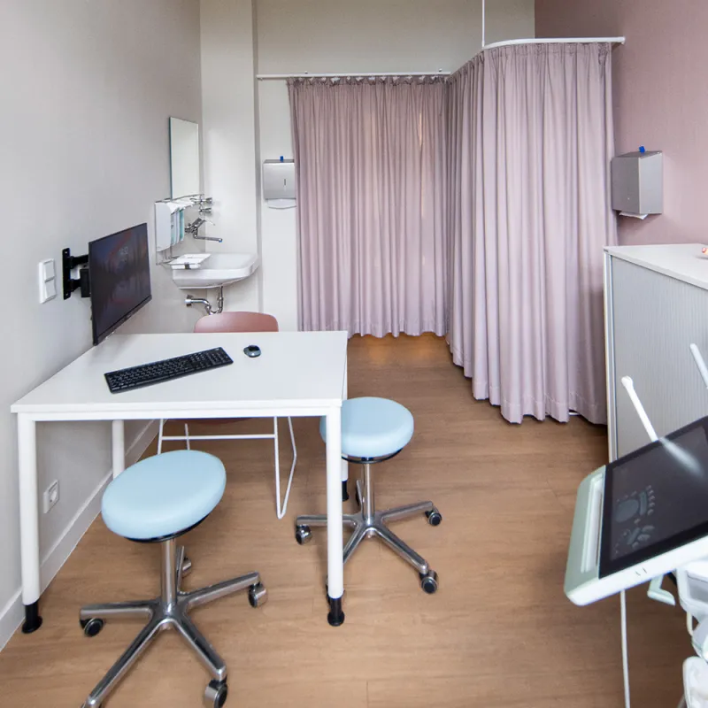 Bürgerhospital – Helles Untersuchungszimmer mit moderner Liege, Holzoptik-Boden und rosa Akzentwand.
