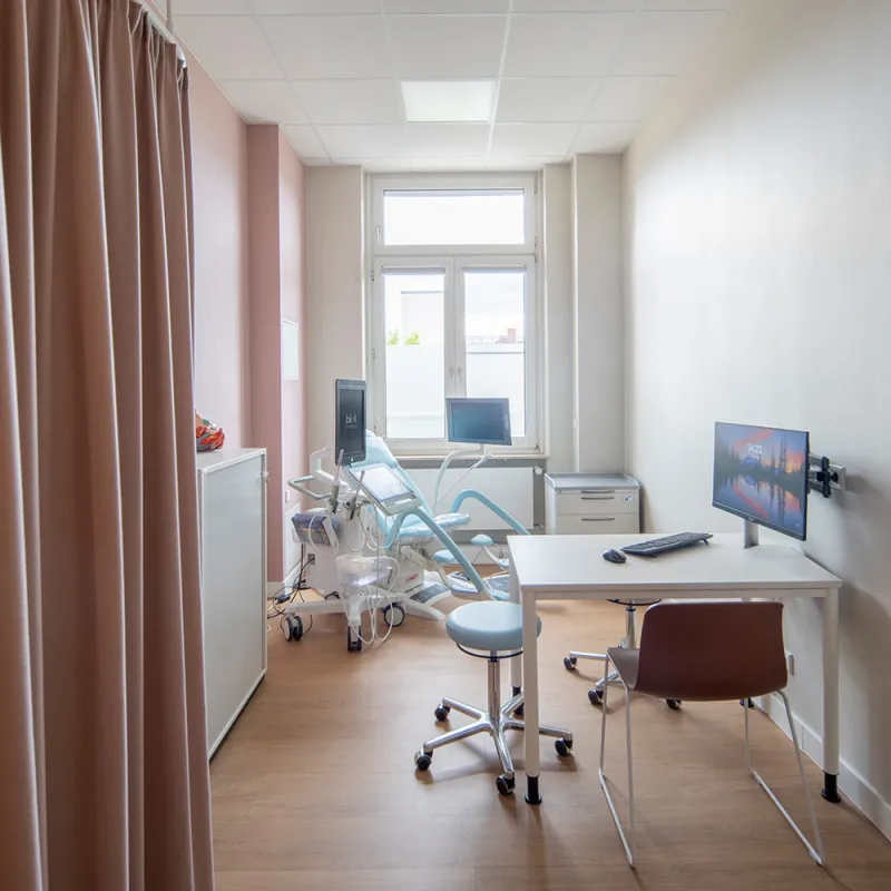 Bürgerhospital – Helles Untersuchungszimmer mit moderner Liege, Holzoptik-Boden und rosa Akzentwand.